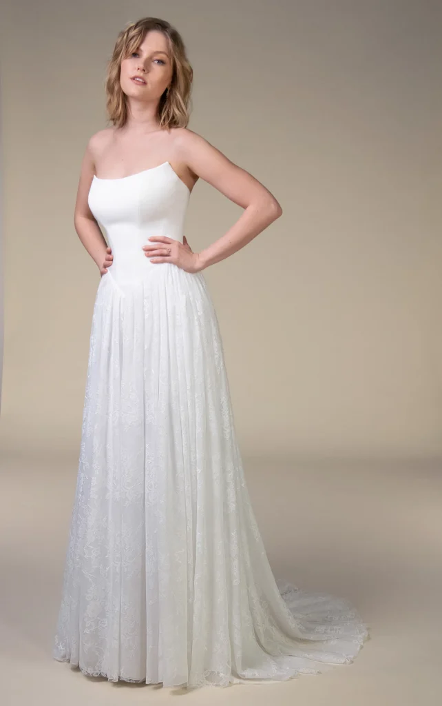 Model wearing the All Who Wander East wedding dress with a strapless sculpted bodice, dropped waist and flowing floral lace A-line skirt, available at The Bridal Collection in Denver, Colorado.