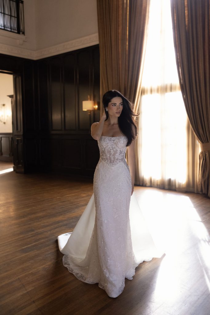 Model wearing the Essense of Australia D4567 wedding dress with a square neckline, sheer lace corset bodice, sparkling sheath silhouette and detachable overskirt