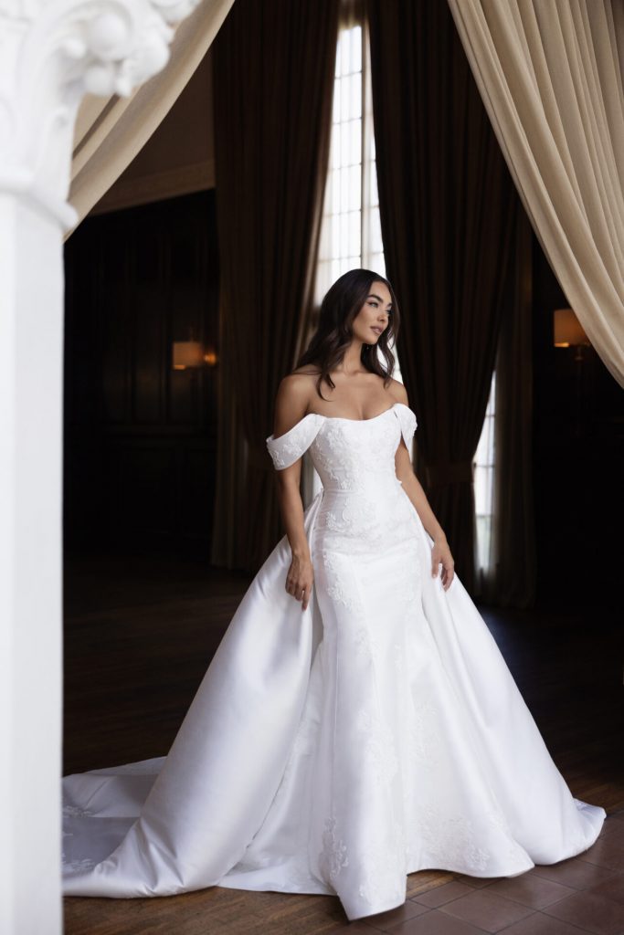 Model wearing the Essense of Australia D4493 wedding dress with an off-the-shoulder neckline, matte lace fit-and-flare silhouette and dramatic overskirt.