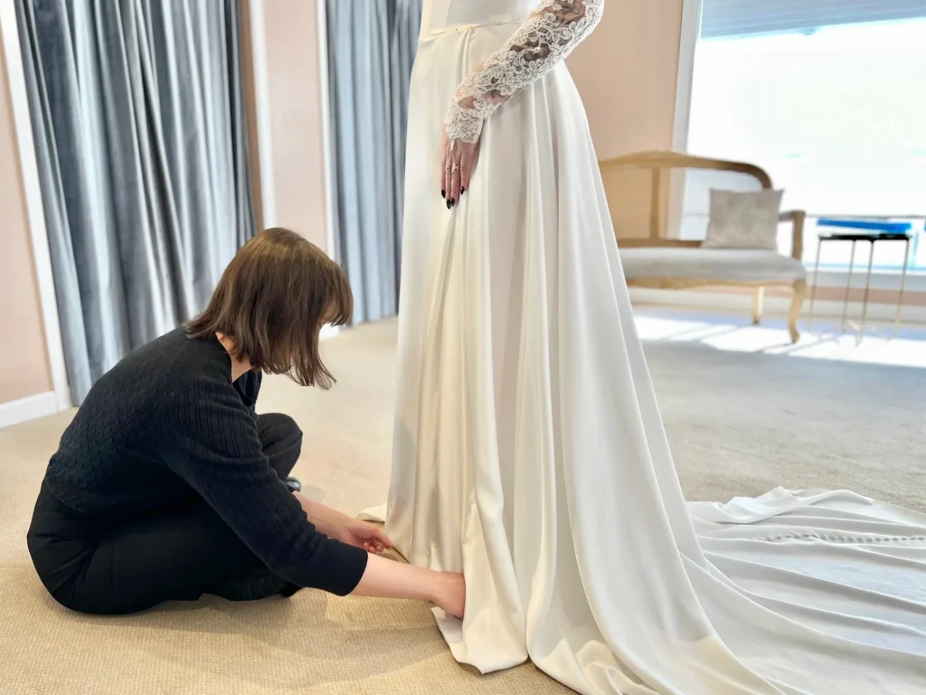Bride standing on pedestal while seamstress pins the hem of her wedding dress.