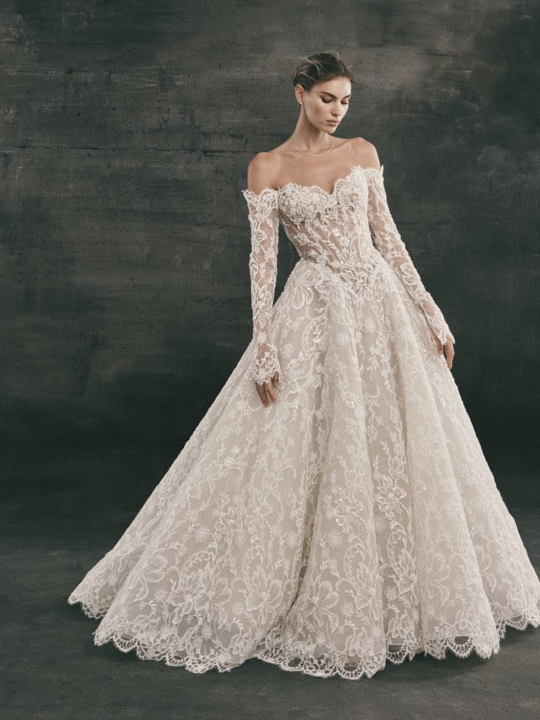 An elegant Nicole + Felicia off-the-shoulder, long-sleeve ballgown wedding dress made of intricate ivory lace with a sheer, textured bodice and scalloped edges, set against a dark grey textured studio background.
