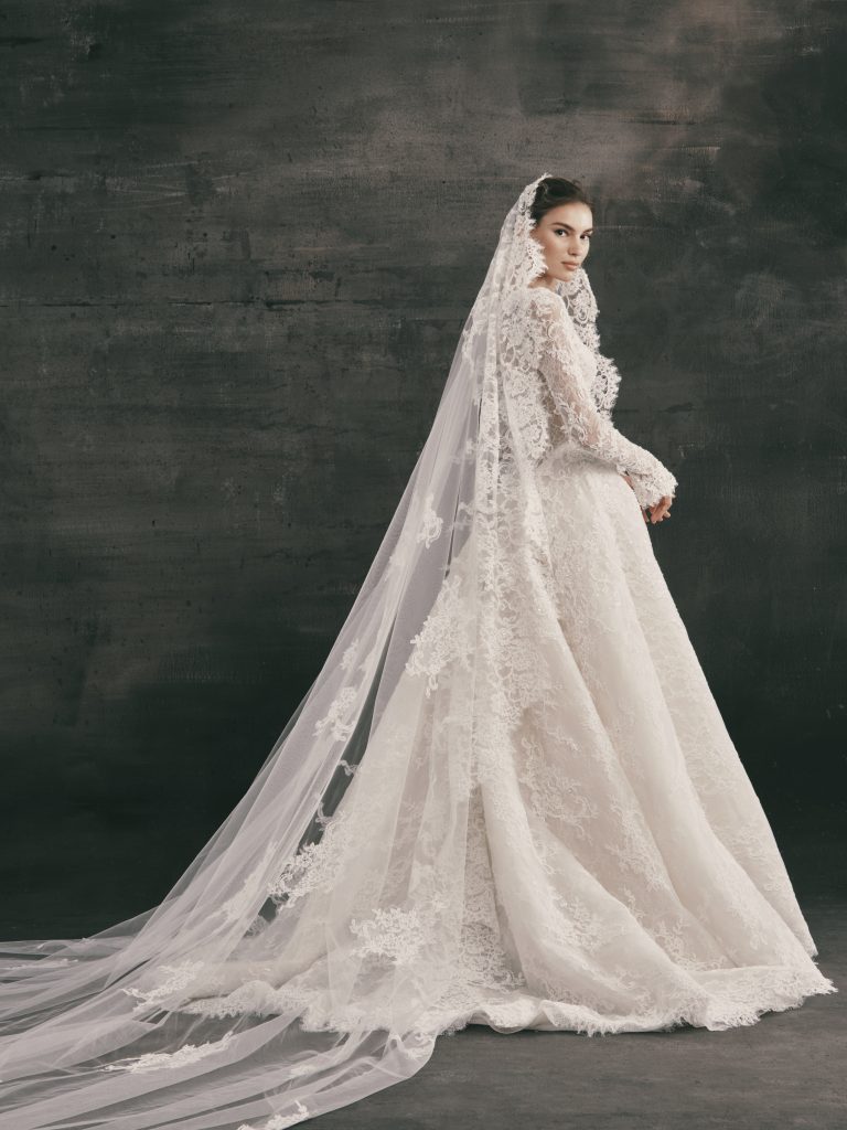Side profile of a Nicole + Felicia Luxe long-sleeve lace ballgown wedding dress paired with a matching cathedral-length lace-trimmed veil, featuring intricate all-over floral embroidery and a dramatic train against a dark grey studio backdrop.