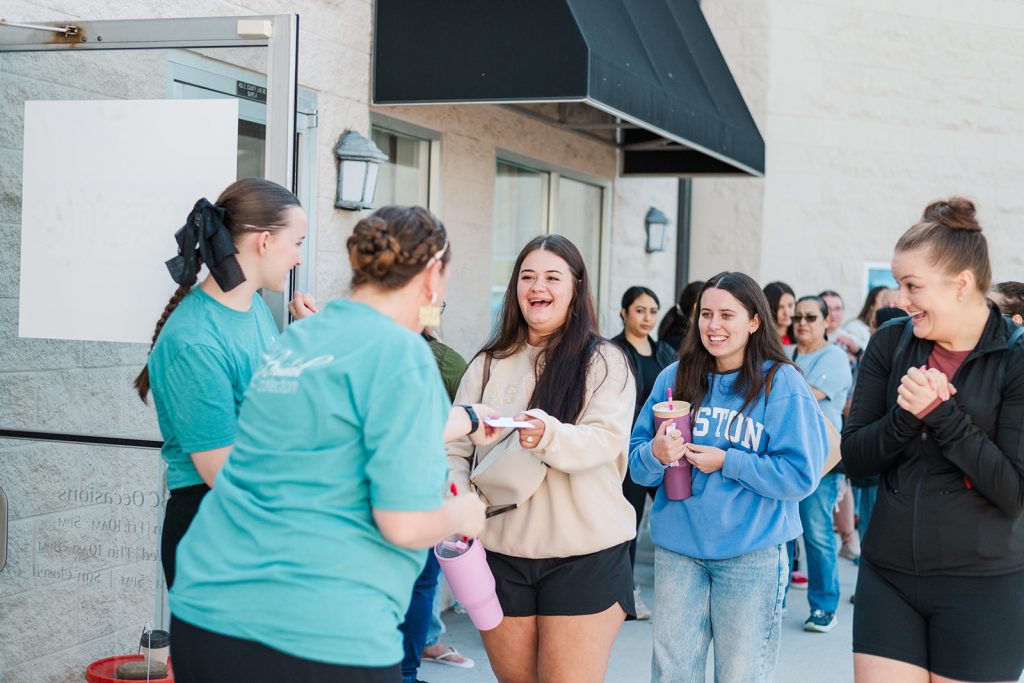 A long line of women stands outside a building with a beige brick exterior under a black awning. On the far left, a staff member in a teal shirt holds a microphone and addresses the crowd. The women in line look on with anticipation, some holding drinks, water bottles, or small party favors.