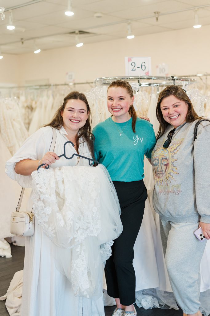 Three smiling women pose together inside a bridal boutique with racks of white wedding gowns in the background. The woman on the left holds a white lace bridal veil on a black hanger. The woman in the center wears a teal shirt with "yes!" scripted on the pocket, and the woman on the right wears a grey sweatshirt and grey sweatpants.