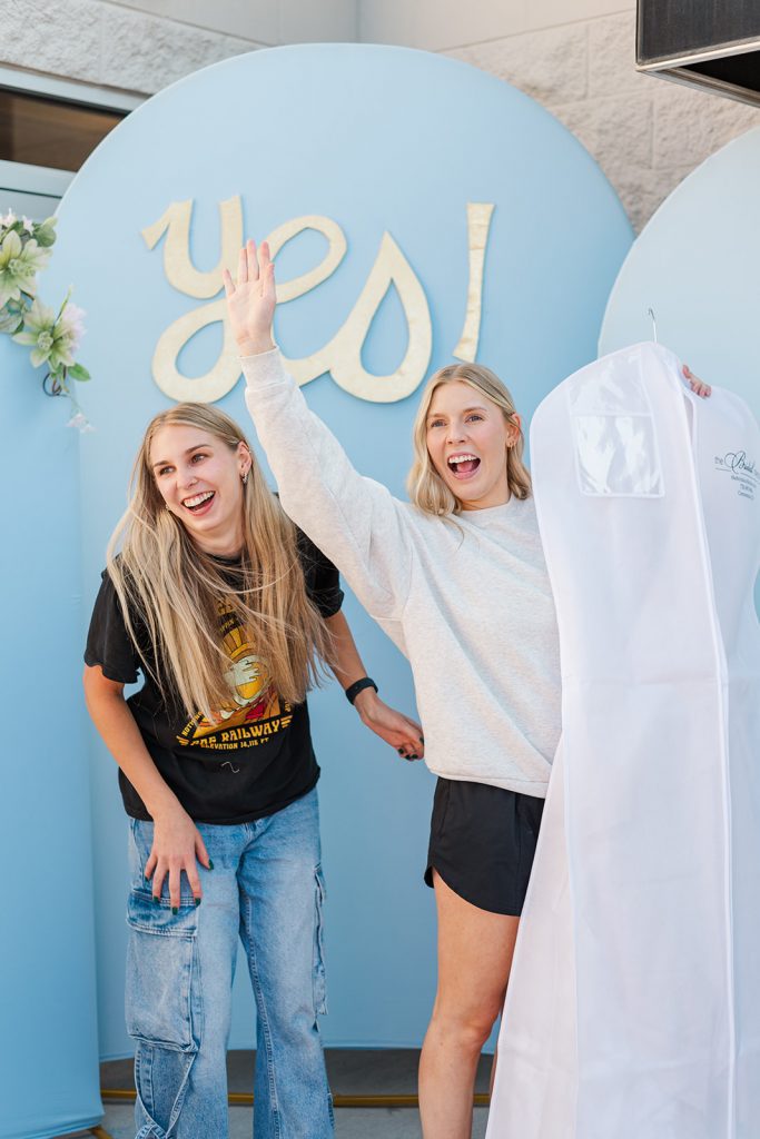 Two joyful women celebrate in front of a light blue backdrop featuring the word "yes!" in gold lettering. The woman on the right wears a grey sweatshirt, laughs excitedly, raises one arm in the air, and holds up a white zippered garment bag containing a dress. The woman on the left wears a black graphic T-shirt and wide-leg jeans, laughing along with her.