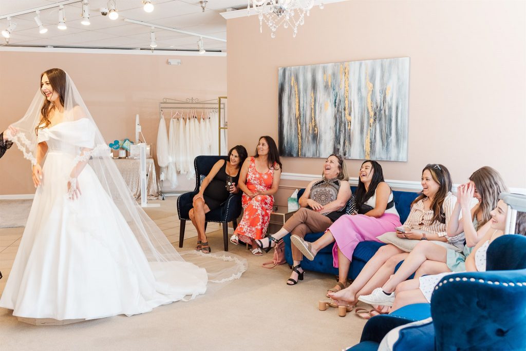 A smiling bride-to-be models an elegant, off-the-shoulder white wedding gown and a long, lace-trimmed veil inside a bridal boutique. She stands on a viewing pedestal while a group of seven women sit on blue velvet couches and armchairs watching her. The guests are laughing and reacting with joy, creating a warm, celebratory atmosphere under bright studio lighting.