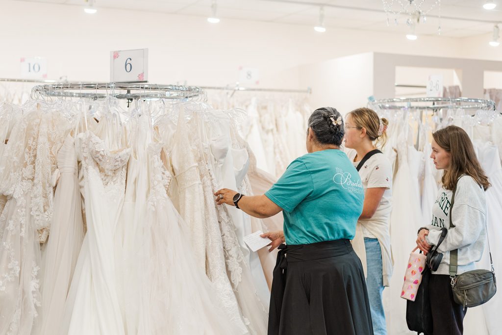 A staff member in a teal shirt helps shoppers browse racks of wedding dresses hanging on round displays during National Bridal Sale Day at The Bridal Collection.