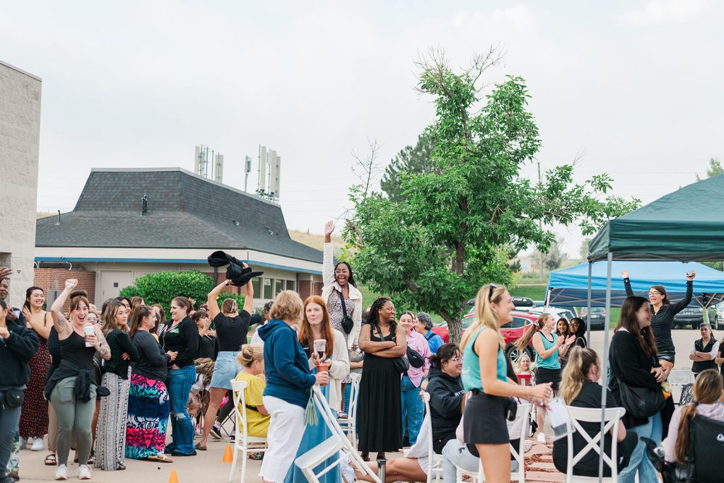 An excited crowd of brides gathering outdoors near event tents during National Bridal Sale Day, with several attendees cheering, waving their hands, and standing together outside of The Bridal Collection.